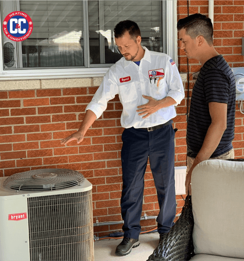 An HVAC technician explains an outdoor air conditioning unit to a homeowner on a brick patio. Both are standing beside the unit. | C & C Heating & Air Conditioning