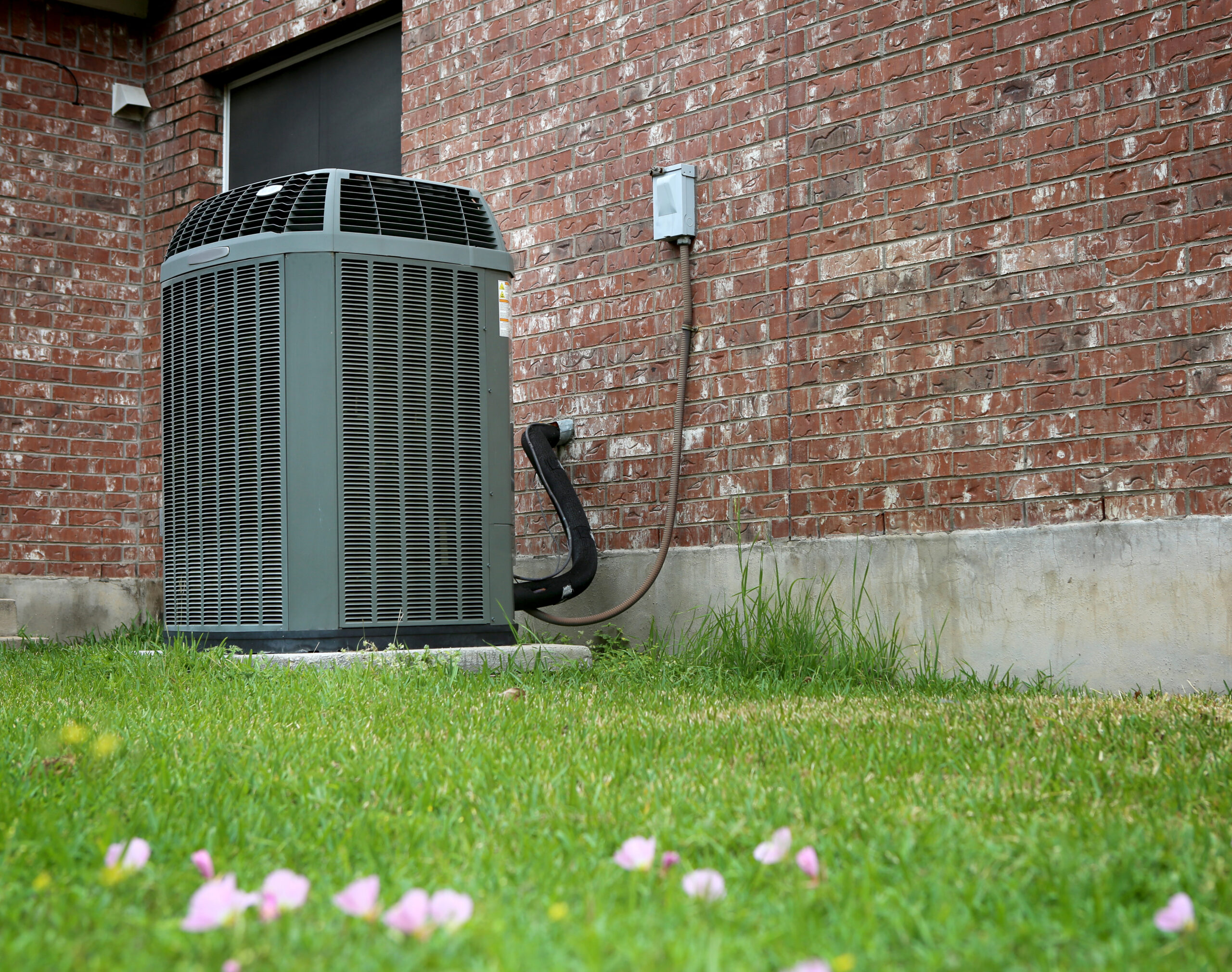 An outdoor AC unit with higher SEER ratings sits next to a brick house, connected by electrical wiring, with grass and scattered flower petals in the foreground. | C & C Heating & Air Conditioning