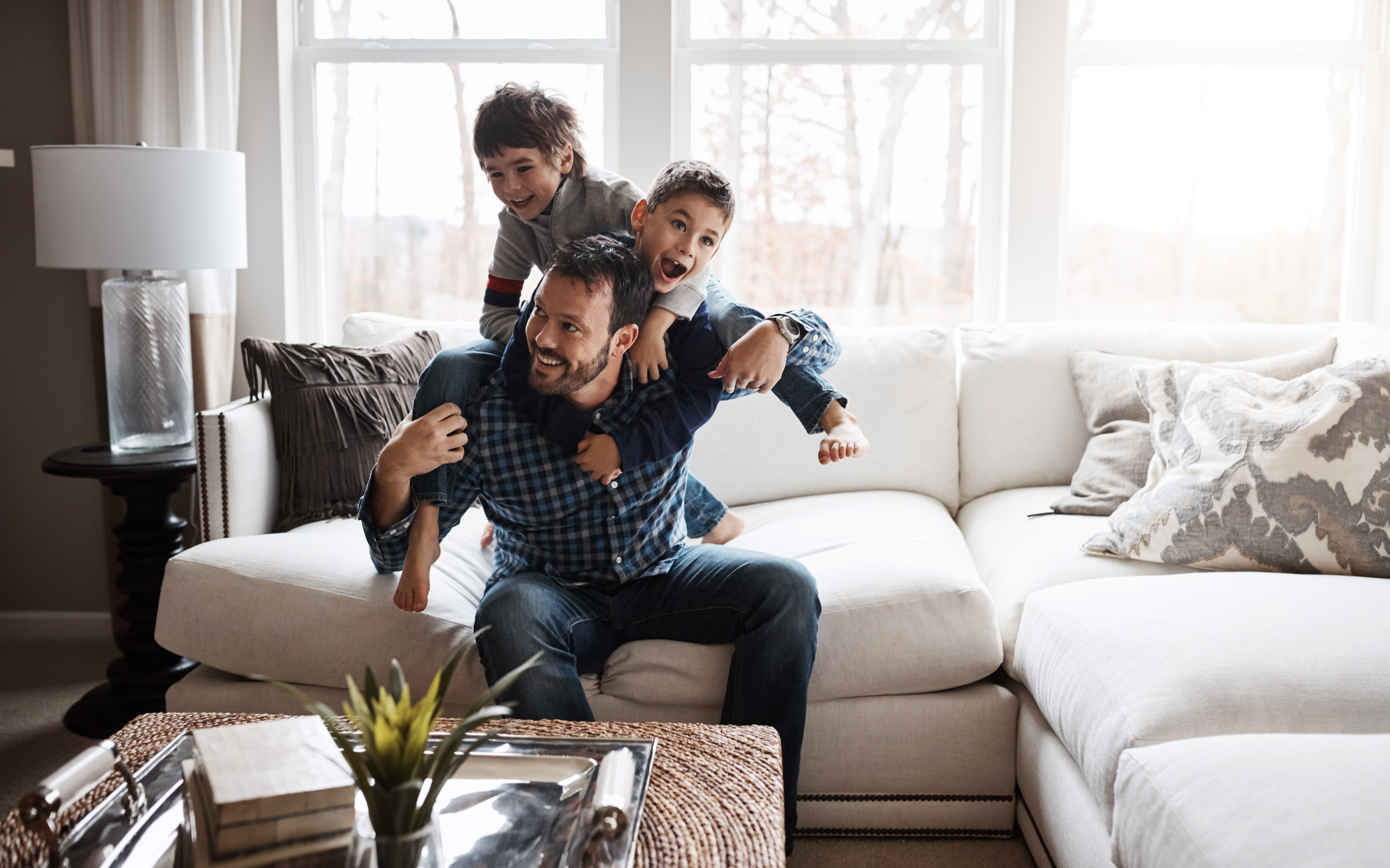An adult sits on a white sofa in a living room with excellent indoor air quality, smiling as two children climb on his back. | C & C Heating & Air Conditioning