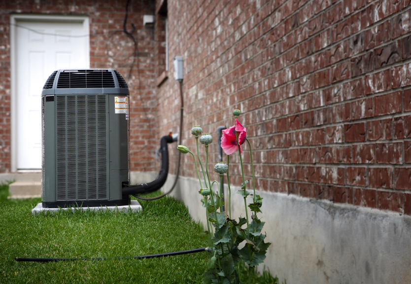 An outdoor air conditioning unit sits on a lawn beside a brick house, with blooming pink poppy flowers nearby—a perfect example of summer HVAC essentials for keeping your home cool and comfortable. | C & C Heating & Air Conditioning