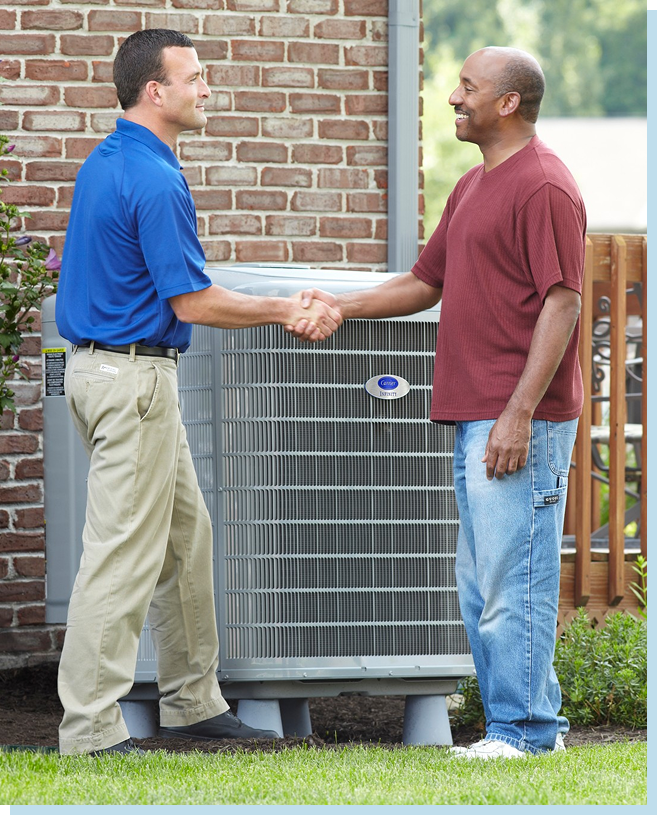 A technician in uniform shakes hands with a man in casual clothes next to an outdoor air conditioning unit by a brick wall. | C & C Heating & Air Conditioning