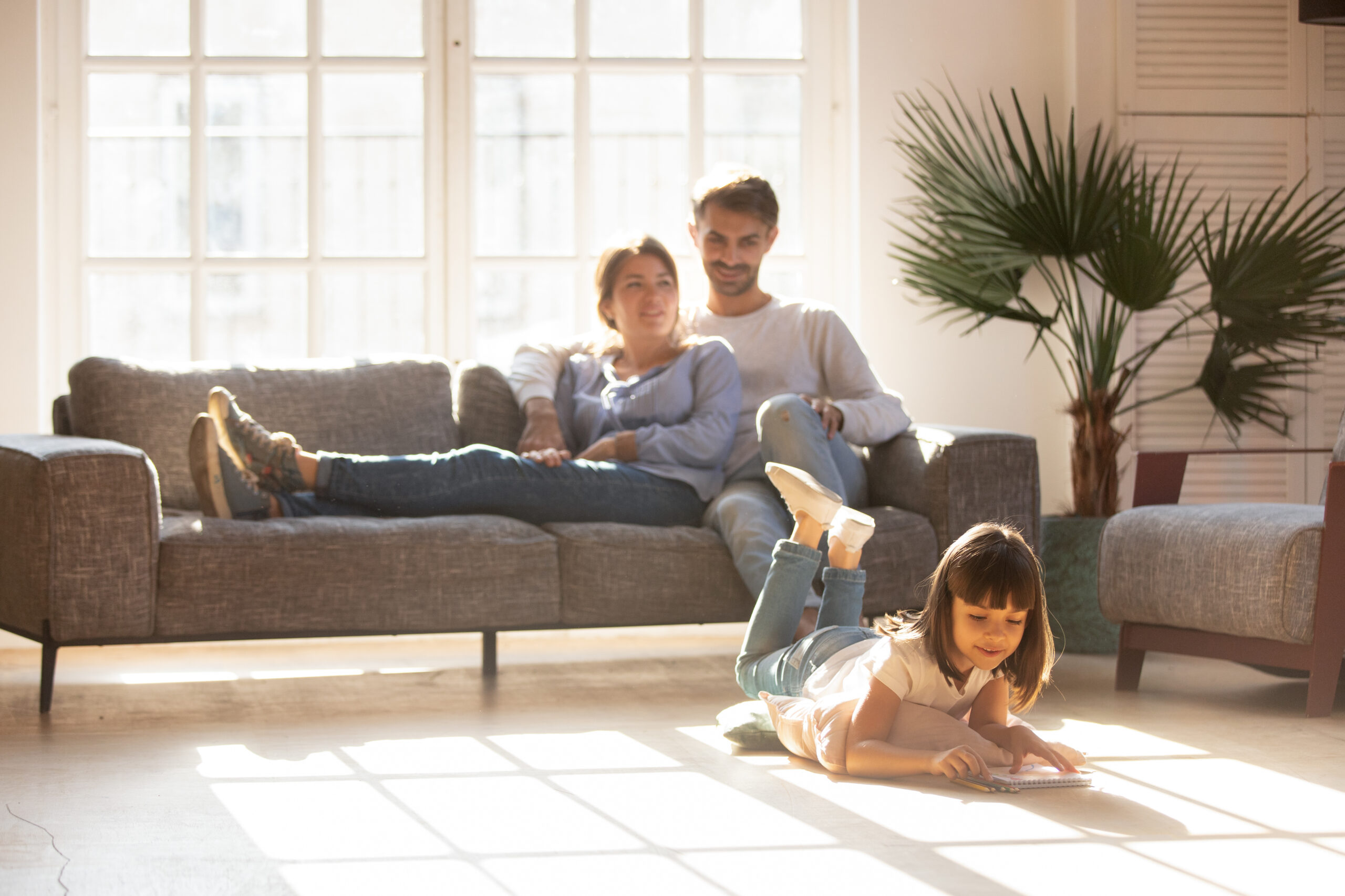 A young girl draws on the floor while two adults relax on a sofa in a bright living room with large windows and indoor plants, which can help improve indoor air quality—a useful Detroit home tip. | C & C Heating & Air Conditioning