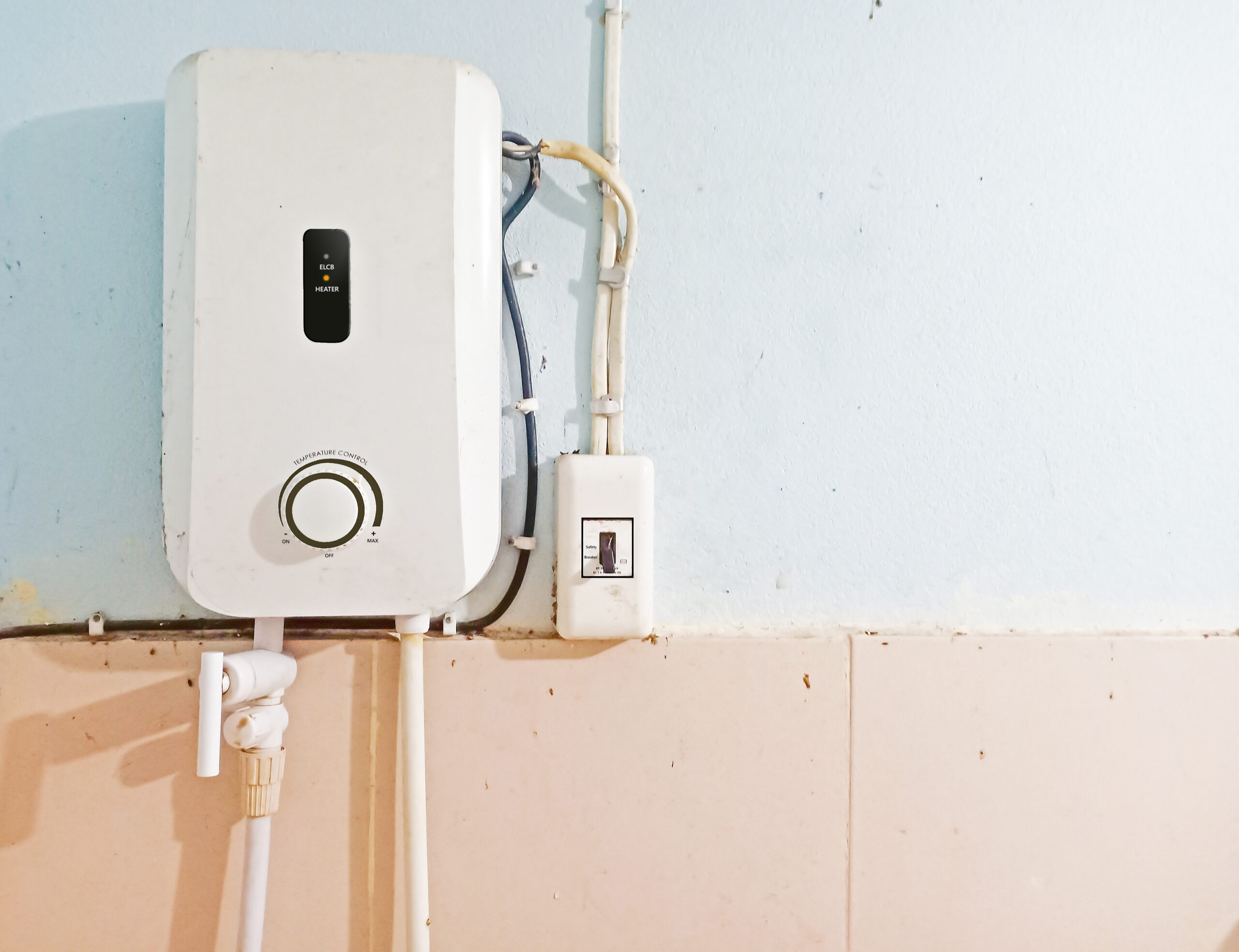 A white electric water heater mounted on a light blue wall, featuring connected pipes and a nearby electrical switch—showing the difference between traditional water heaters and tankless water heaters in design and installation. | C & C Heating & Air Conditioning