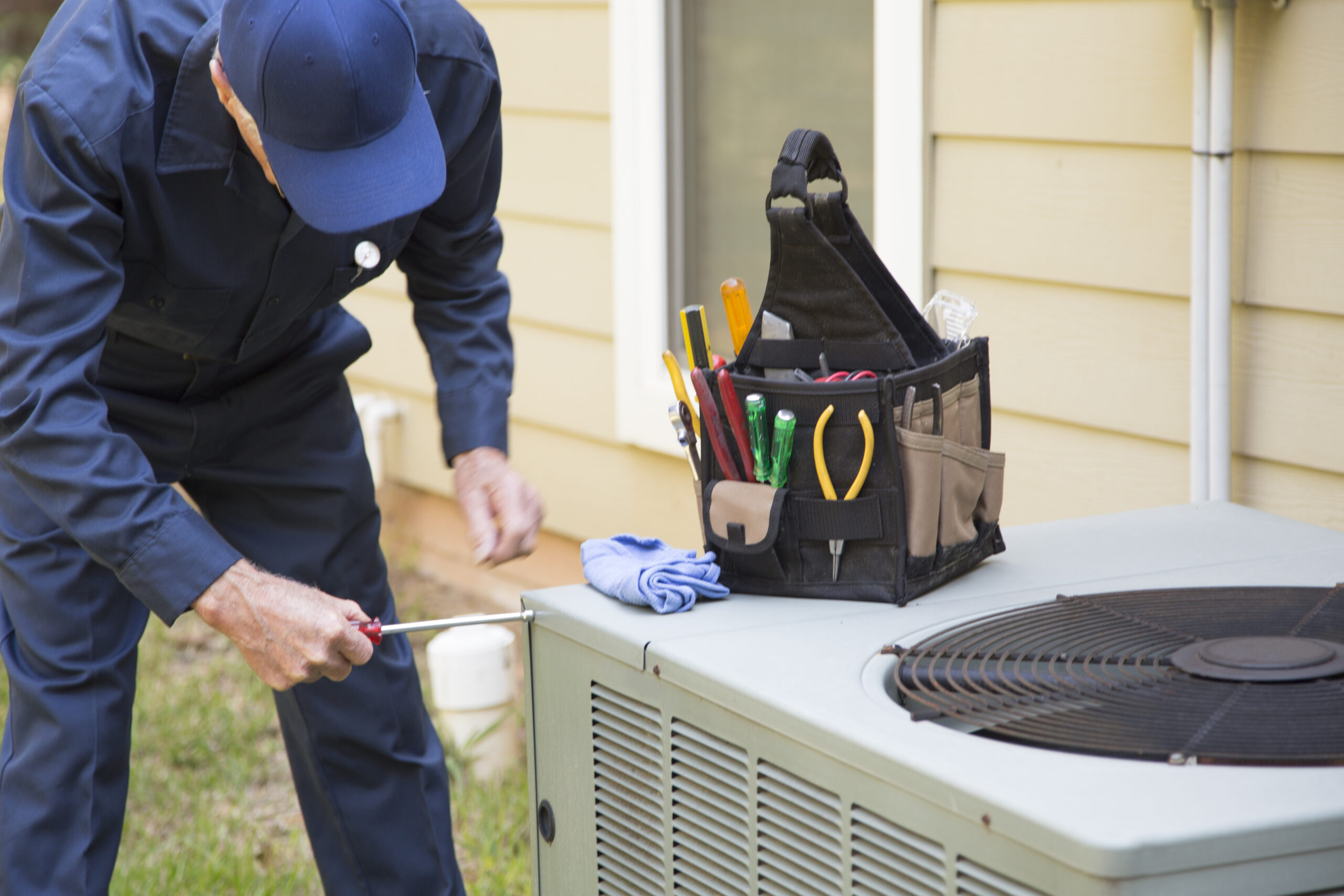 A technician in uniform from a trusted HVAC company uses a screwdriver to service an outdoor air conditioning unit next to a house, keeping cool air flowing; a tool bag and cloth rest on the unit. | C & C Heating & Air Conditioning
