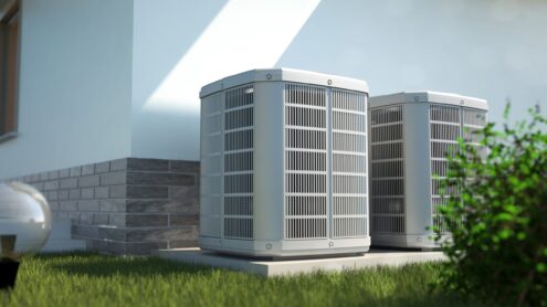 Two outdoor central air conditioning units are installed on concrete pads next to the exterior wall of a house, with grass and a small bush nearby, allowing for easy comparison with heat pumps in a similar setup. | C & C Heating & Air Conditioning