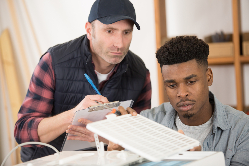 Two men, possibly engaged in apprenticeships or training from trade schools, inspect and discuss a white object—one taking notes on a clipboard while the other examines it closely. | C & C Heating & Air Conditioning