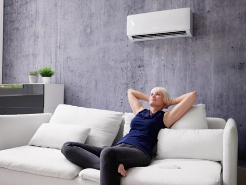 A woman relaxes on a white sofa in a modern living room with a wall-mounted air conditioner above her, enjoying Spring indoor air and knowing she can breathe easier. | C & C Heating & Air Conditioning
