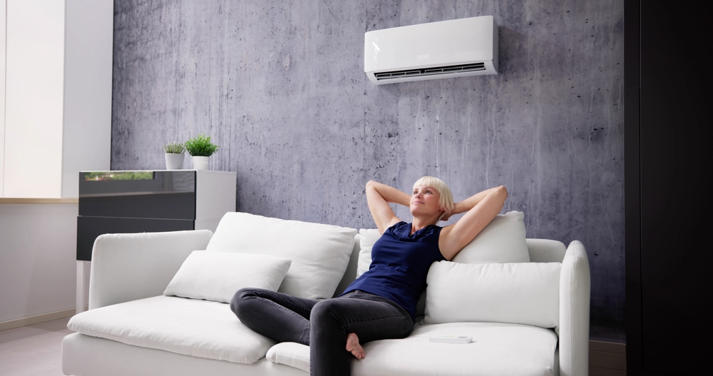 A woman relaxes on a white sofa in a modern living room with a wall-mounted air conditioner above her, enjoying Spring indoor air and knowing she can breathe easier. | C & C Heating & Air Conditioning