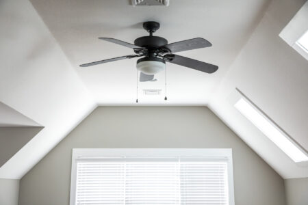A ceiling fan with four blades and a light fixture is mounted on the ceiling of a bright room with skylights and a window with closed blinds, helping you save energy and lower your energy bill. | C & C Heating & Air Conditioning