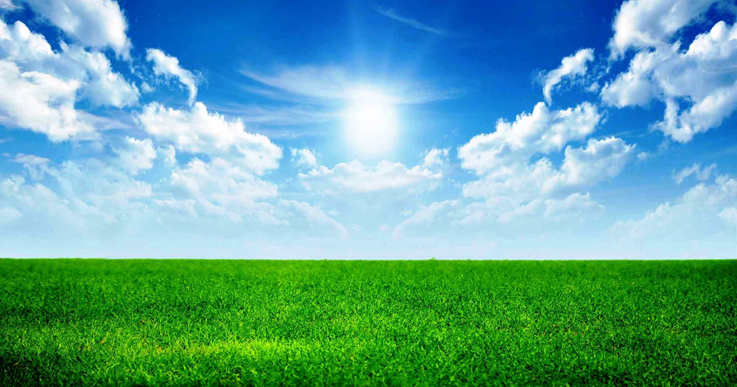 A bright sun shines in a blue sky with scattered clouds above a wide, green grassy field, illustrating the positive environmental impact of using clean energy solutions like heat pumps. | C & C Heating & Air Conditioning