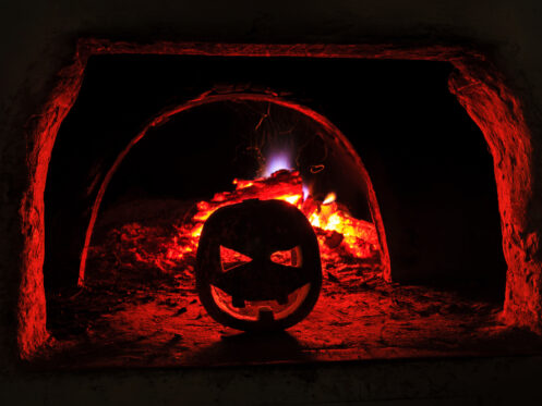 A jack-o'-lantern sits in front of a fire inside a dark brick oven, its red-orange light illuminating the scene—a perfect reminder to treat your furnace with the same warm care this season. | C & C Heating & Air Conditioning