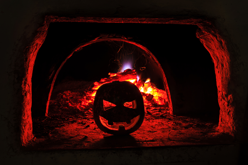 A jack-o'-lantern sits in front of a fire inside a dark brick oven, its red-orange light illuminating the scene—a perfect reminder to treat your furnace with the same warm care this season. | C & C Heating & Air Conditioning