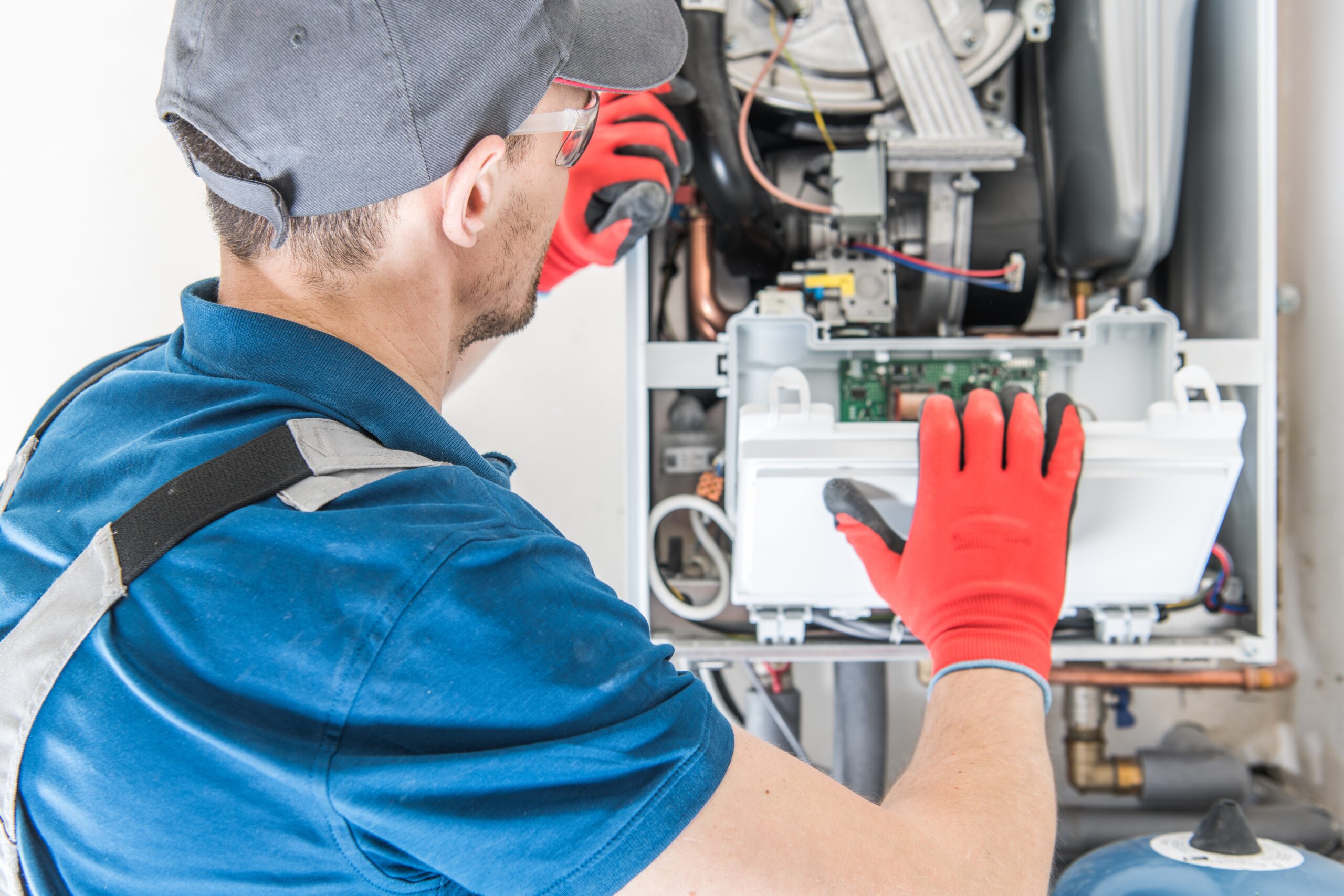 A technician wearing red gloves and a cap repairs an open boiler, inspecting internal components and wiring, possibly diagnosing furnace problems or addressing a banging noise inside the unit. | C & C Heating & Air Conditioning