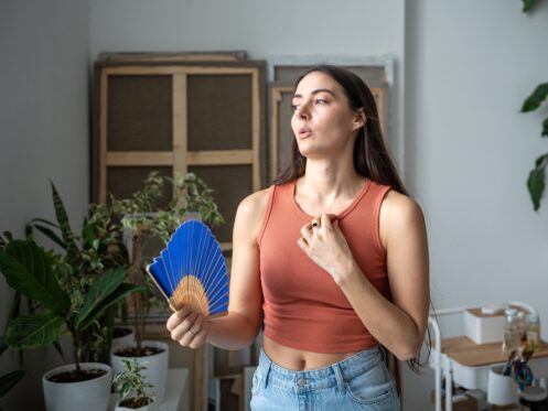 A woman in a sleeveless top fans herself with a blue hand fan while standing indoors among potted plants, highlighting the importance of indoor humidity for comfort and air quality. | C & C Heating & Air Conditioning