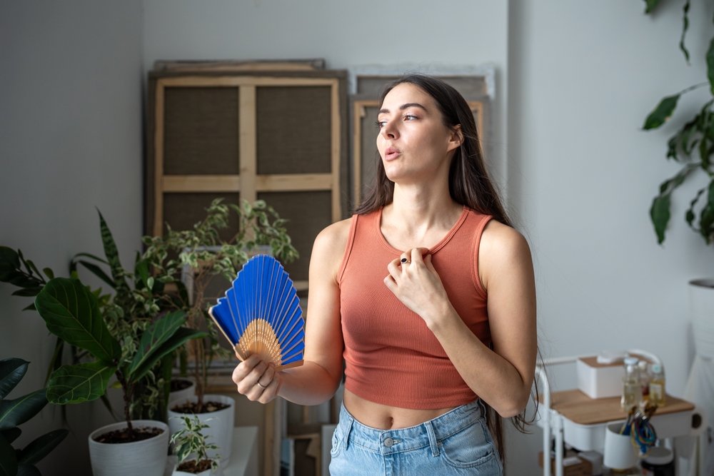 A woman in a sleeveless top fans herself with a blue hand fan while standing indoors among potted plants, highlighting the importance of indoor humidity for comfort and air quality. | C & C Heating & Air Conditioning