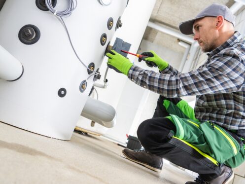 A worker in a plaid shirt and green pants adjusts a control box on a large water heater, ensuring it’s ready to meet summer demand. | C & C Heating & Air Conditioning