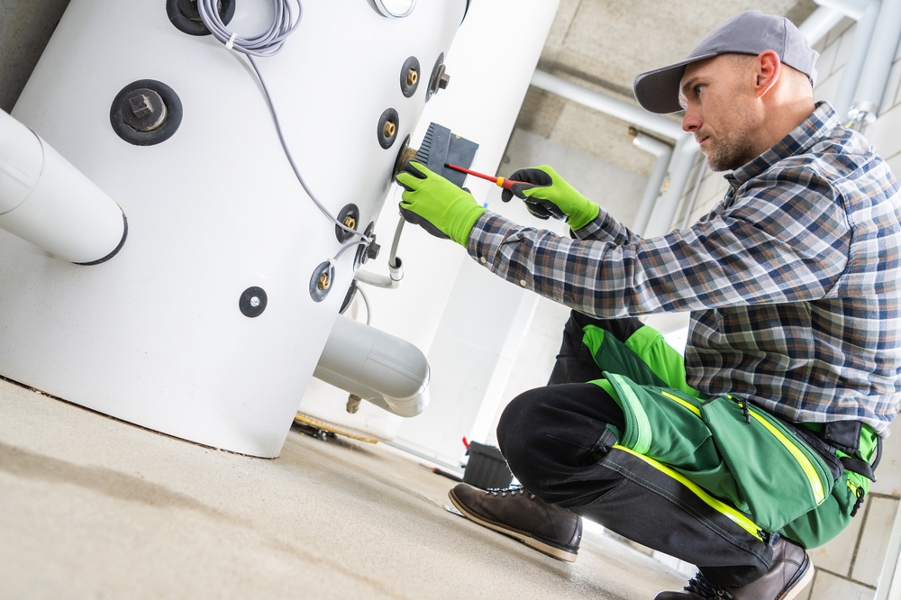 A worker in a plaid shirt and green pants adjusts a control box on a large water heater, ensuring it’s ready to meet summer demand. | C & C Heating & Air Conditioning