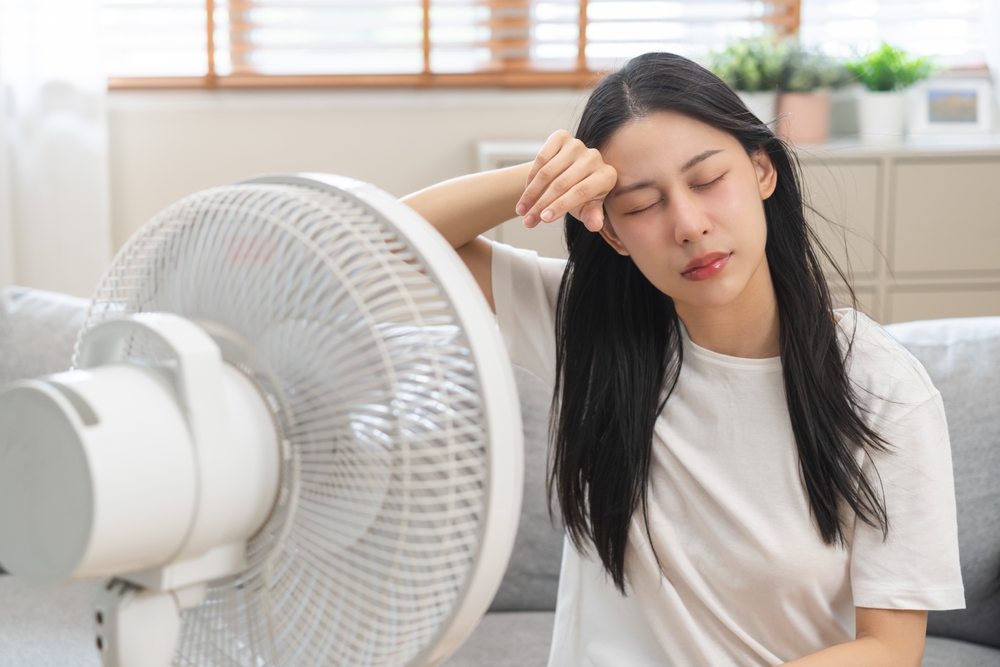 A woman sits on a couch in front of a fan, wiping her forehead and appearing overheated in a brightly lit living room—clearly wishing she were ready with AC for Summer 2025. | C & C Heating & Air Conditioning