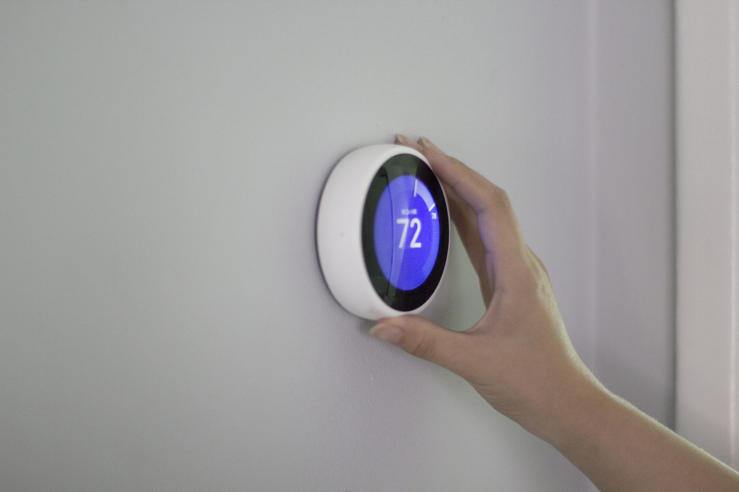 A hand adjusts a smart thermostat mounted on a light gray wall, showcasing one of the key advantages of smart thermostats—easy control and efficient temperature settings in your smart home. | C & C Heating & Air Conditioning