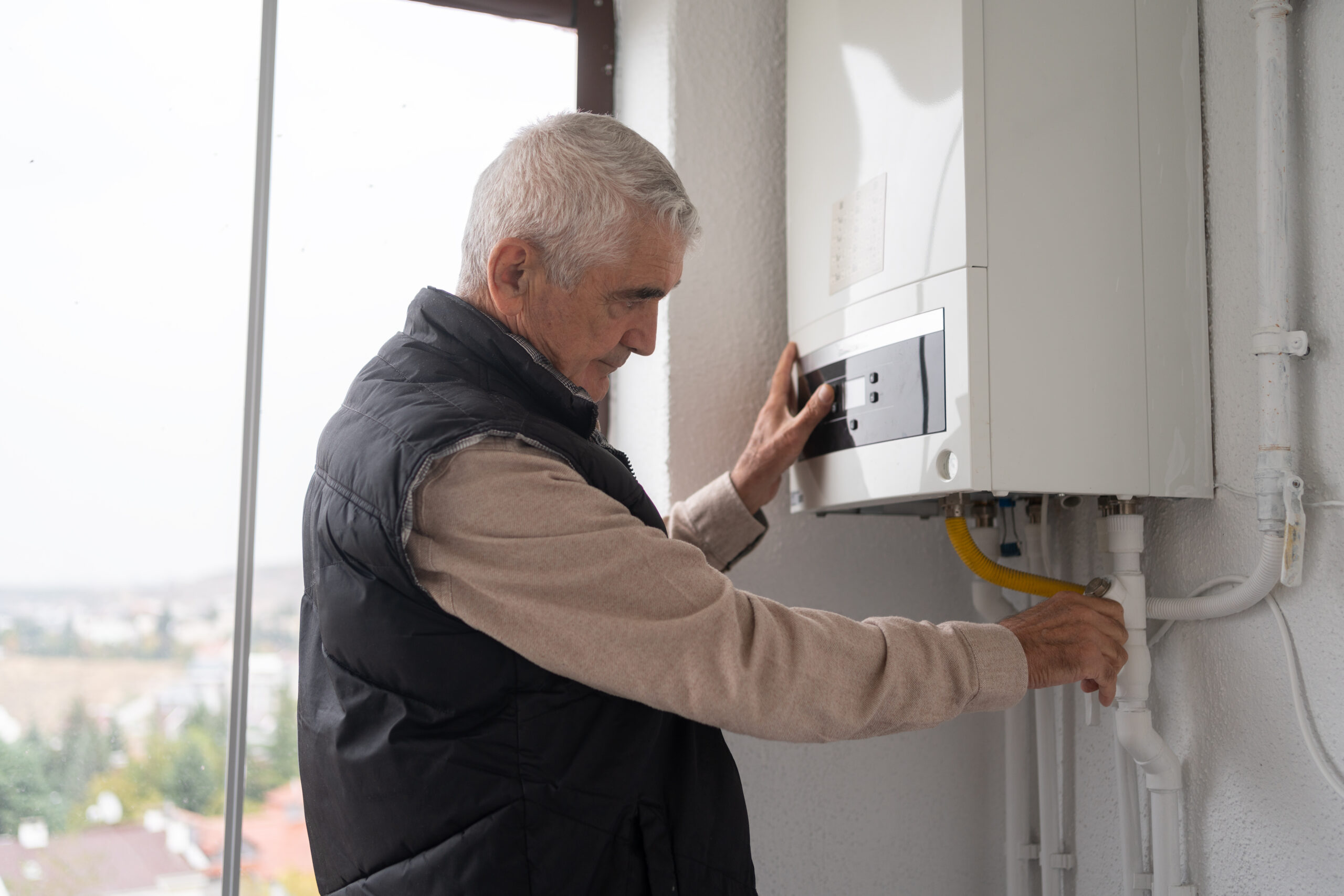 An older man in a vest adjusts a control on a wall-mounted gas boiler, illustrating the difference between traditional water heaters and modern tankless water heaters in a room with a large window. | C & C Heating & Air Conditioning