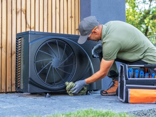 A man wearing gloves and a cap cleans an outdoor HVAC unit with a cloth, focusing on energy efficiency; a tool bag with various tools is next to him on the ground. | C & C Heating & Air Conditioning