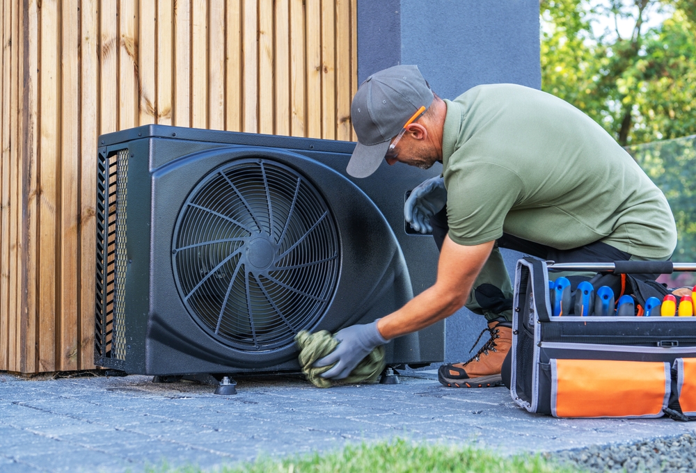 A man wearing gloves and a cap cleans an outdoor HVAC unit with a cloth, focusing on energy efficiency; a tool bag with various tools is next to him on the ground. | C & C Heating & Air Conditioning