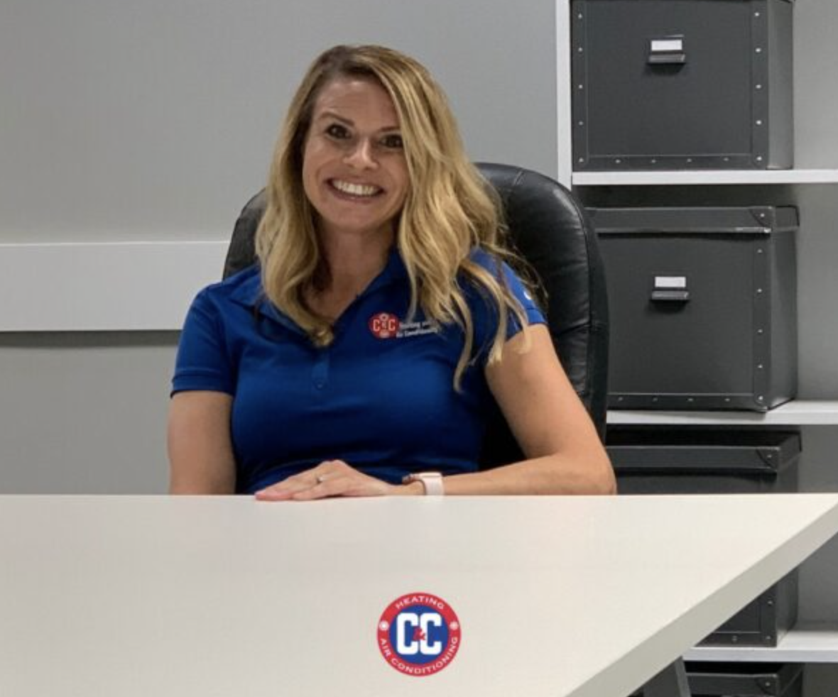 A woman with long blonde hair, wearing a blue polo shirt with a CC logo, sits at a white desk in an office with gray storage boxes—representing Women in the Trades and their impact on Trades History. | C & C Heating & Air Conditioning